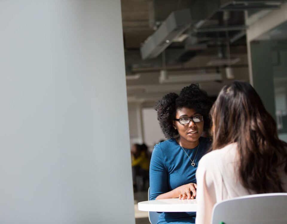 O Que é Recursos Humanos e Qual a Sua Importância nas Empresas? Two women engaged in a discussion in a modern office environment, highlighting communication and interaction.