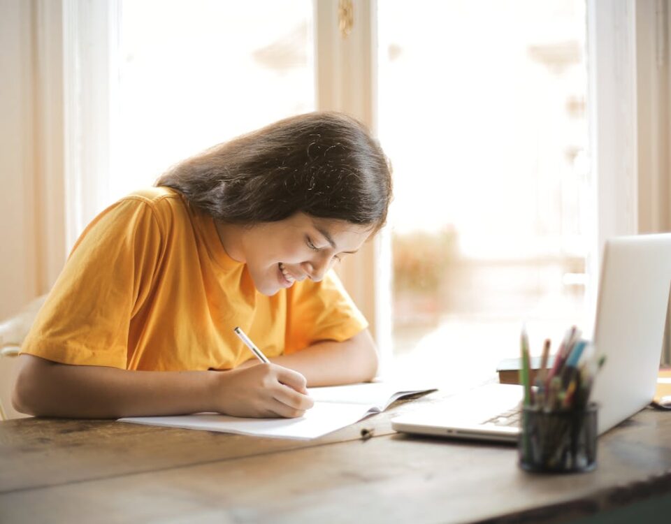 Algoritmos ajudam a criar métodos de estudo personalizados Young woman studying with a laptop and notepad at home, smiling and focused.