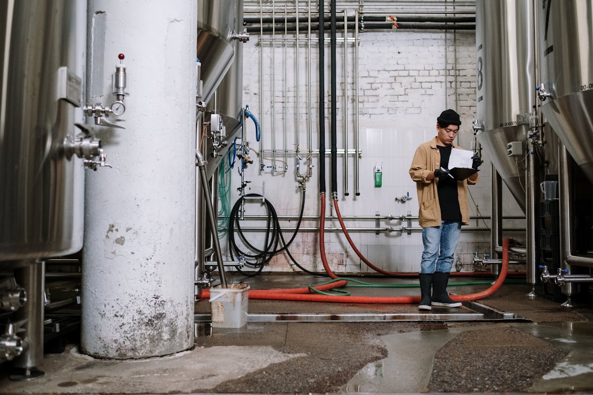 A brewery worker inspects equipment in a brewing facility, surrounded by fermentation tanks.