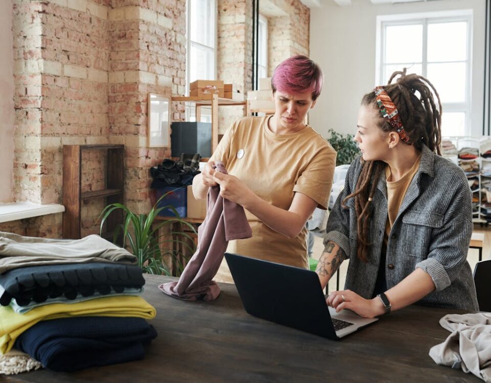 Como a inteligência artificial impacta a moda infantil Two women sorting clothing donations using a laptop in a bright urban setting.