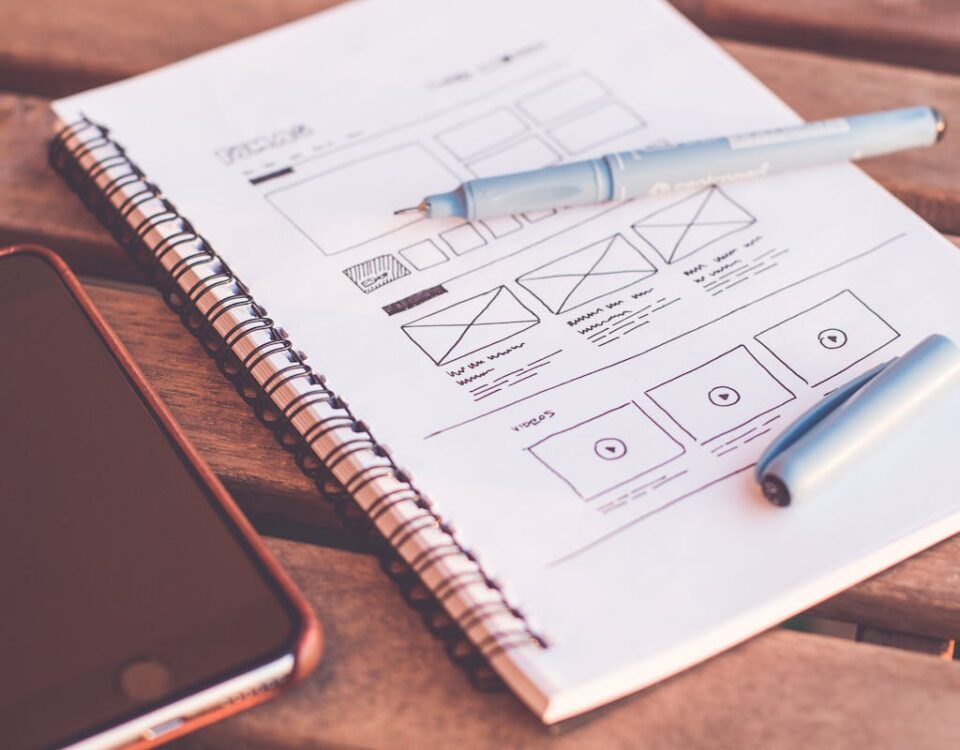 Como os frameworks web aceleram o desenvolvimento de sites Close-up of a notebook with wireframe sketches and a smartphone on a wooden desk.