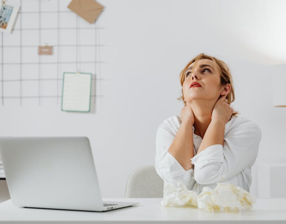 Inteligência artificial pode prever desconforto físico? A stressed woman in white long sleeves sits at a desk with a laptop, looking upwards in an office.
