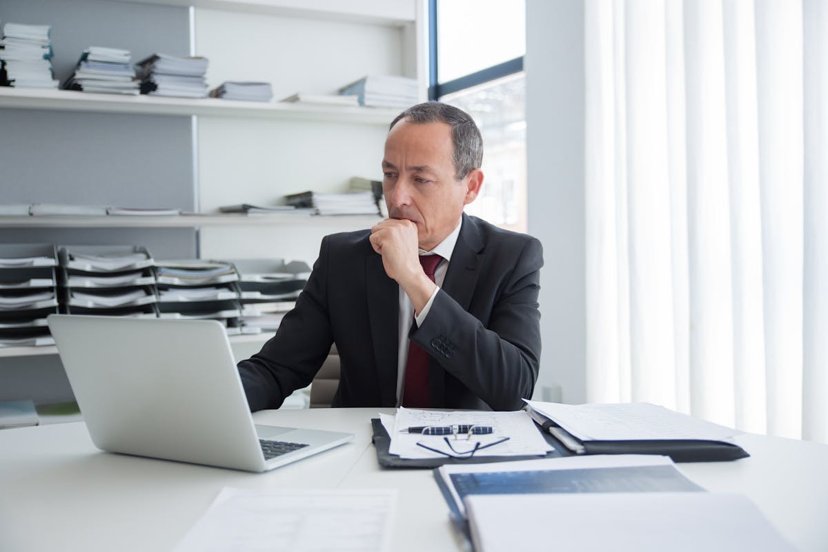 Mature business executive thoughtful at desk with laptop and documents in modern office setting.