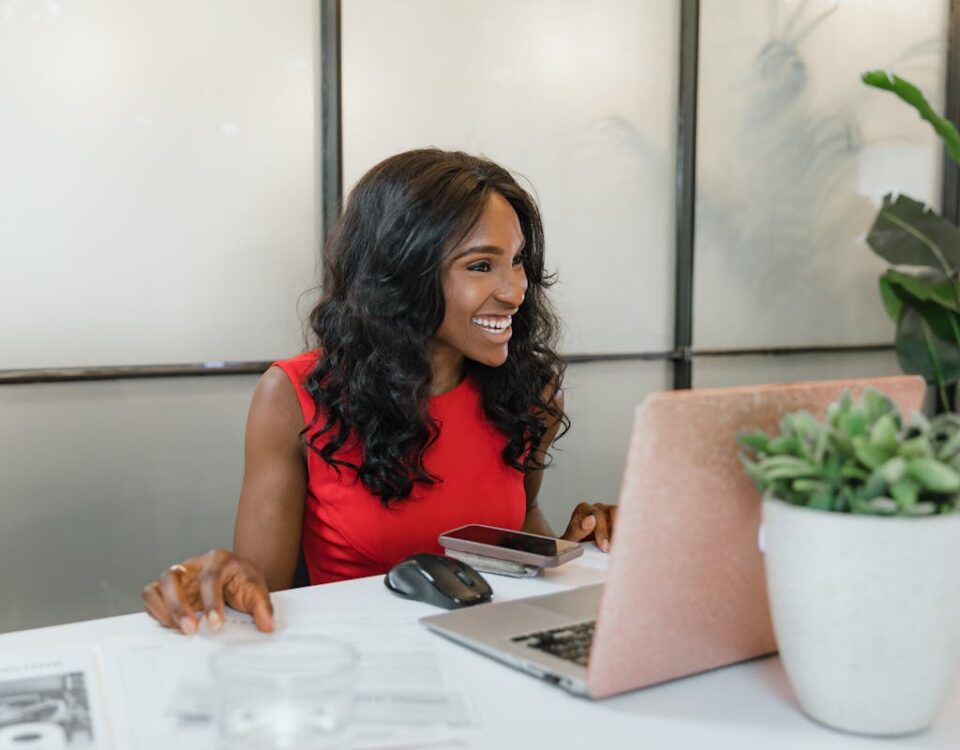 Algoritmos que melhoram a eficiência administrativa Professional black woman smiling at desk using laptop and smartphone in office.