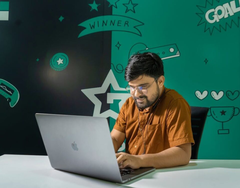 A man with a beard and glasses smiles while gaming on a laptop indoors, decorated with gaming graphics.