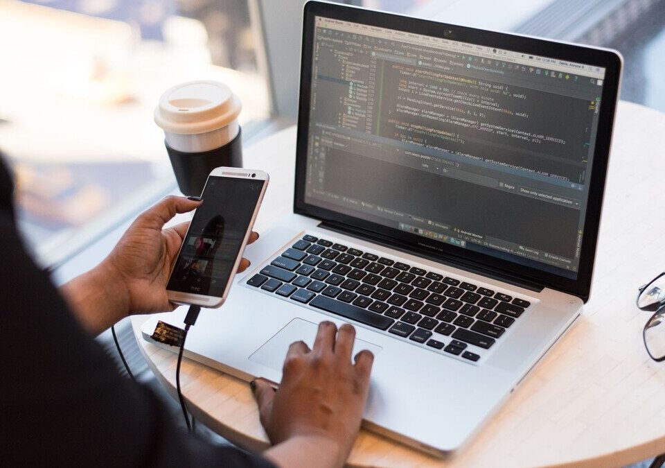 Pessoa segurando um celular enquanto usa um notebook com código na tela