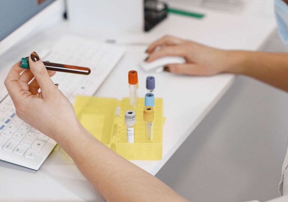 Profissional de laboratório conferindo tubos de amostra ao lado de um computador
